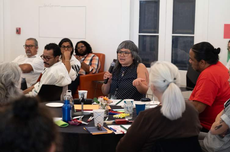 Lan Tuazon, a woman with Filipino heritage, long dark hair laced with gray and asymmetrical bangs, gestures with one hand and speaks into a microphone from a table, and various other people at her table and other tables listen attentively.