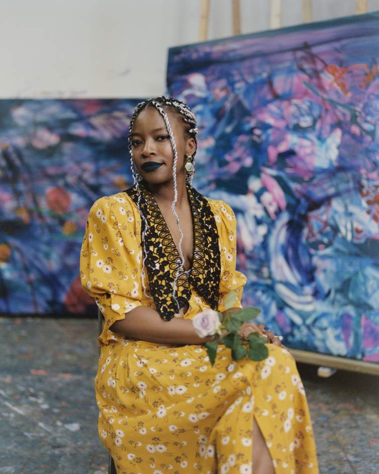 Jadé Fadojutimi looks directly at the camera serenely, holding a rose in her lap. She is a Black woman with medium skintone, long braided hair plaited with black and white strands. She wears black lipstick and a yellow floral dress, behind her are two large abstract paintings.