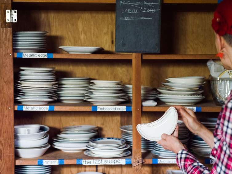 Julie Green holds a small ceramic dish in her hand, facing a wooden shelf holding dozens of varied small dishes. The shelves are labeled Metallic edge, Embossed edge, patterns, and the last supper.
