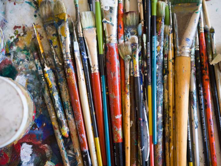 About 20 paintbrushes of various sizes and shapes are clustered on a paint splattered table.