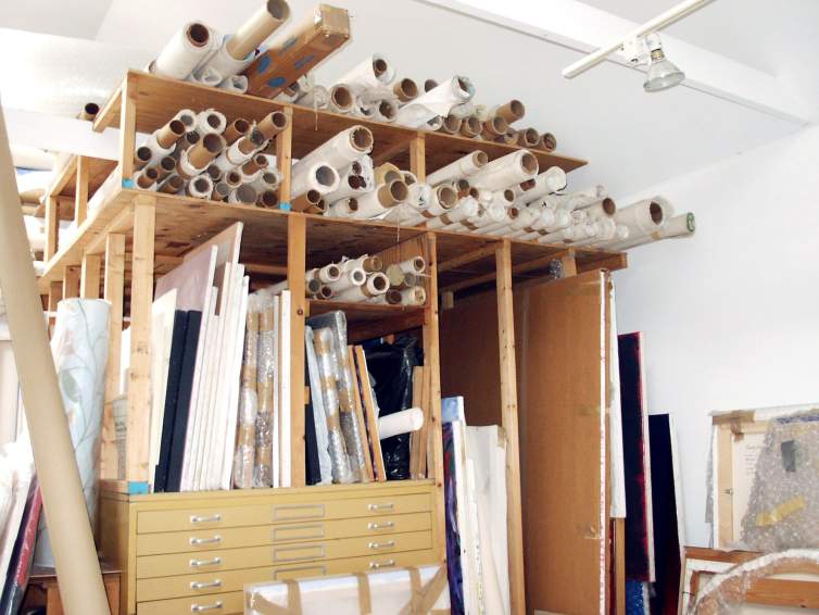 In an art studio stands a large artwork storage rack, which holds at the top dozens of rolled canvases and papers, and below, groups of vertically stored stretched canvases on top of a flat file.