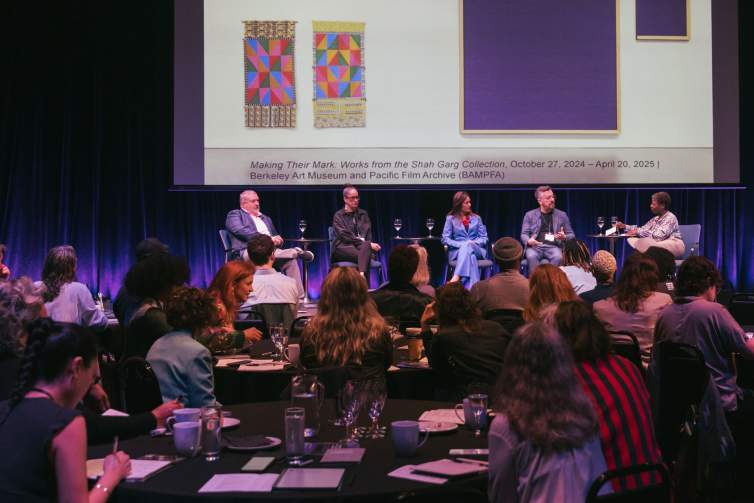 In front of a seated crowd, a panel of speakers sits in chairs in front of a projection of artwork. To the far right, Thelma Golden, a Black woman with short hair, speaks and gestures to the other men and women seated.