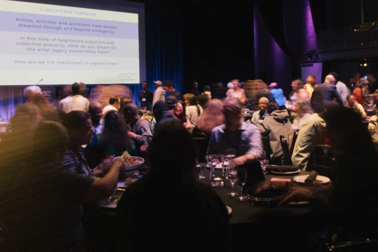 In a darkened room with a projected screen, people move between black tables with some bodies blurred by motion.