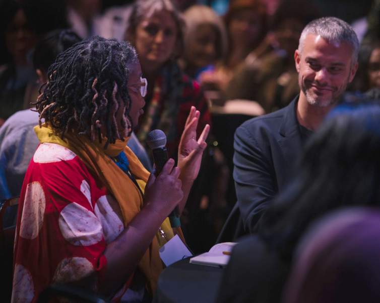 A woman with dark skin tone, short twisted braids, and bright clothing speaks into a microphone at a table in a conference setting. We see a man with medium skin tone and short gray hair smiling at her from across the table.