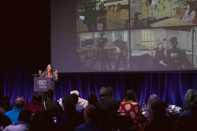An Asian woman with light skin tone presents to a crowd on stage, with a projected image of video stills in the background.