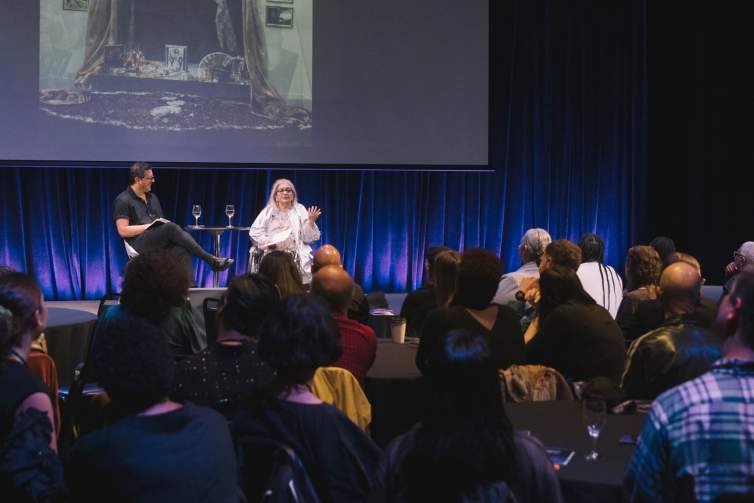 Josh T. Franco and Amalia Mesa-Bains, two people of Mexican American descent, sit on a stage in a room filled with people sitting at round black tables. A screen behind them shows one of Amalia's artworks.