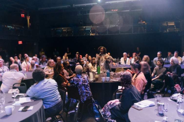 A woman with dark skin tone and curly brown hair stands with a microphone, addressing the group of people seated around her at round tables.