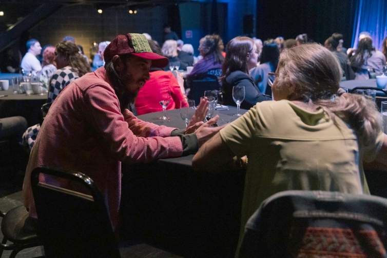A man with light skin and long dark hair, wearing a red trucker cap, sits at a round table with a woman with medium skin tone and gray hair, engaged deeply in conversation.