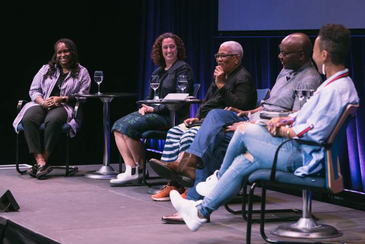 Five people of different ages and skin tones sit on a stage engaged in discussion.