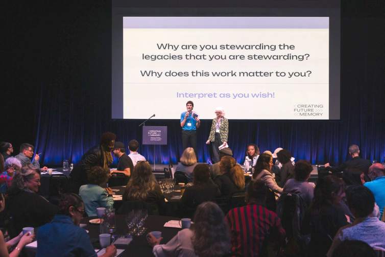 On a stage, two people address the audience at a conference, speaking into microphones. A text slide behind them reads, Why are you stewarding the legacies that you are stewarding? Why does this work matter to you? Interpret as you wish!
