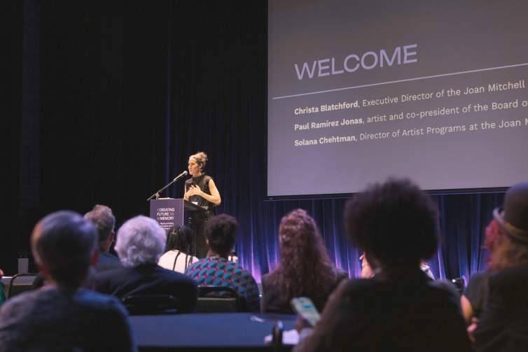 Solana Chehtman, a woman of Hispanic descent with light skin tone and brown hair pulled up in a bun, presents at a podium in front of a crowd seated at tables. On the screen behind her, we see "Welcome" along with her name and the names of other presenters.