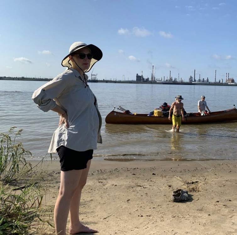 AnnieLaurie standing on land in front of the Mississippi River, with figures in a canoe behind her and Dow Chemical Plant on other side of river