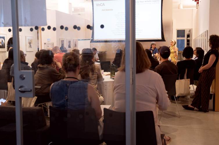 Looking through a glass wall, a group of people at tables watch two white women presenting at the front of a room.