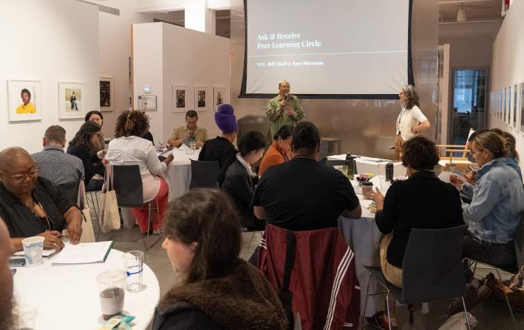 A group of artists sit at round tables in a meeting space as two presenters stand at the front near a projector screen reading reading "Ask & Receive Peer Learning Circle"