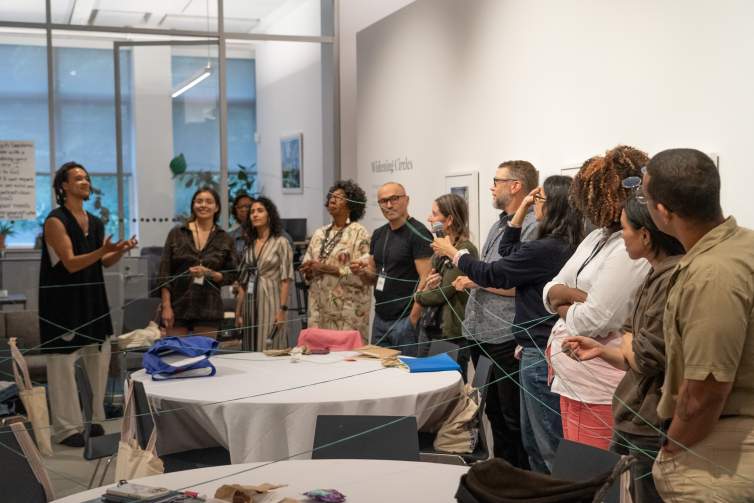 A group of artists stand in a semi-circle, holding green strings connecting them, as Mala Iqbal tosses a green ball of string to Sandy Williams IV.