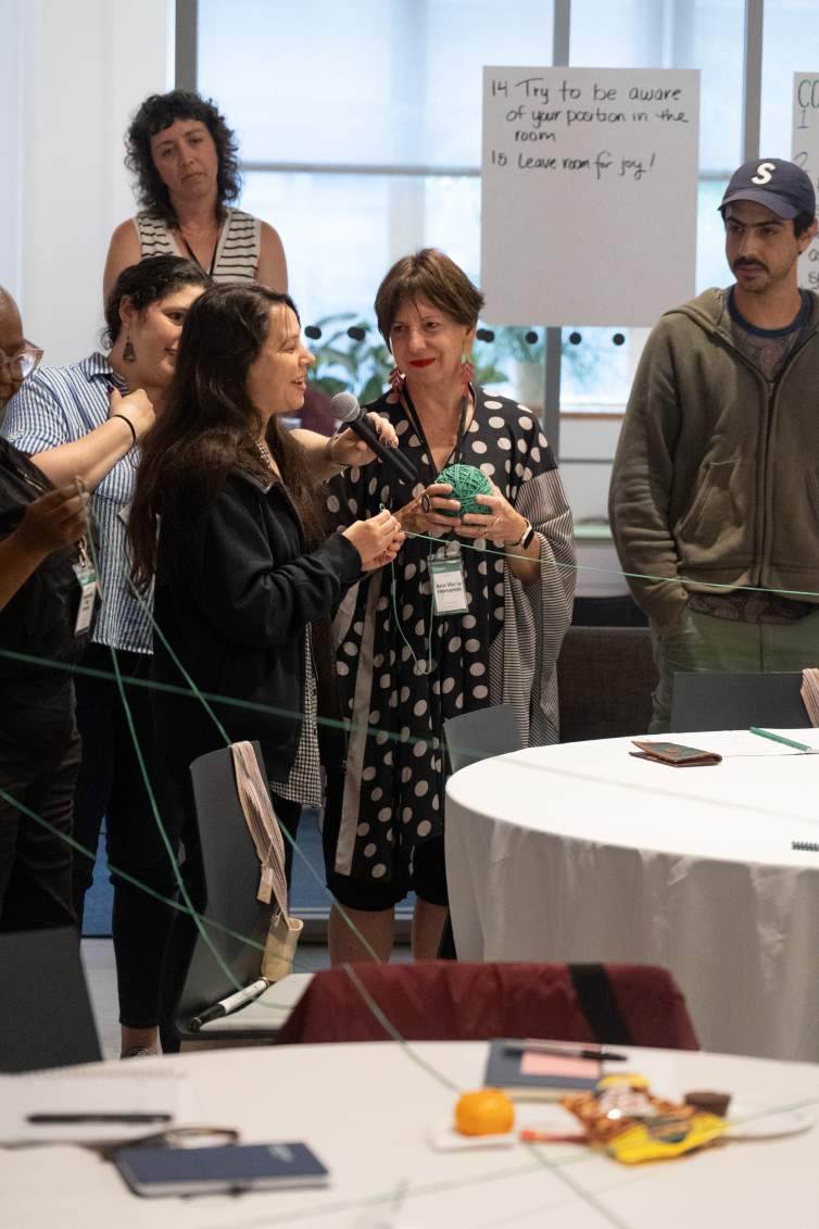 Emilie Louise Gossiaux, a white woman with long brown hair and a black jacket smiles and speaks into a microphone as a group of people look on smiling, holding green strings.