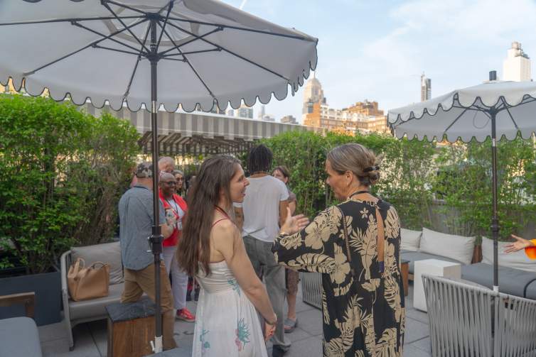 Under scalloped umbrellas on a rooftop with NYC buildings visible behind, several people smile and talk in summery clothing.