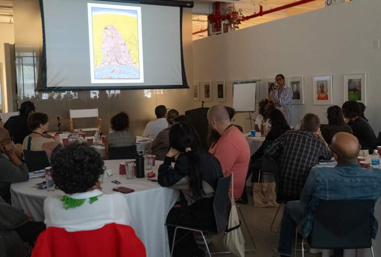 In a darkened room, people at round tables look at a screen with an image of a vertical painting depicts a figure with her head bent downwards in profile, enrobed in a pink hooded cloak. Rupy C. Tut speaks into a microphone, an Indian-American woman with medium skin tone and long dark hair pulled back.
