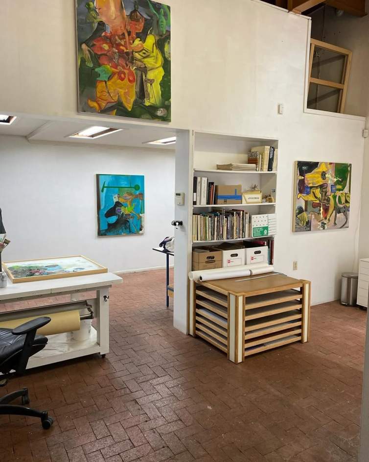 A view of Scott Anderson's painting studio shows a tidy space with ed brick floor, work tables, storage, books, files, and paintings hung on white walls.