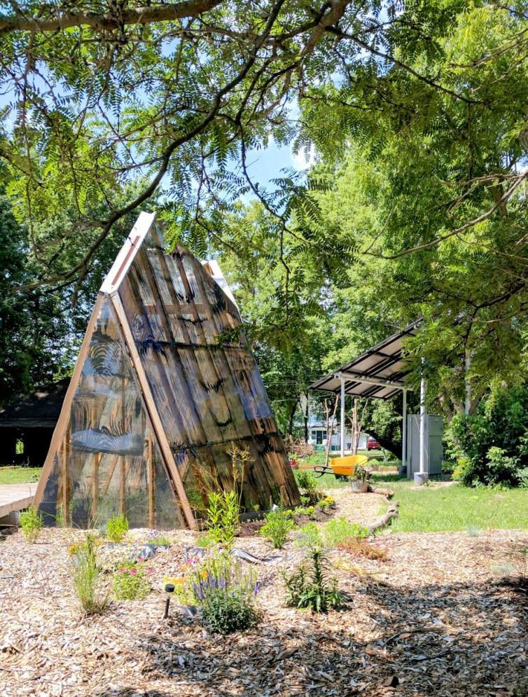 “Shine A Light” is solar-powered triangular pod-like structure, with a structure of cedar boards with a semi translucent sheath with patterns on the surface like oversized wood grain. The structure is set in a garden with cedar mulch and overhanging trees, and a solar array is behind the triangular structure to the right.