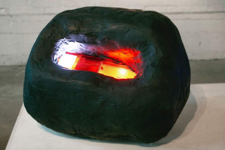 “Joey” is a sculpture of a lit automotive tail light set into a rounded biomorphic black form like a small boulder. The form has a spackled plaster-like texture and a matte finish.