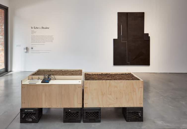 “Broadcast #3” is a sculpture of 4 plywood cubes stacked on top of 2 black milk crates, each filled with soil, and a copper tube protruding from the soil. On a shelf extending from a cube is a cassette player, and a black cable leads to another shelf with a small analog drum machine and other audio equipment with cables.