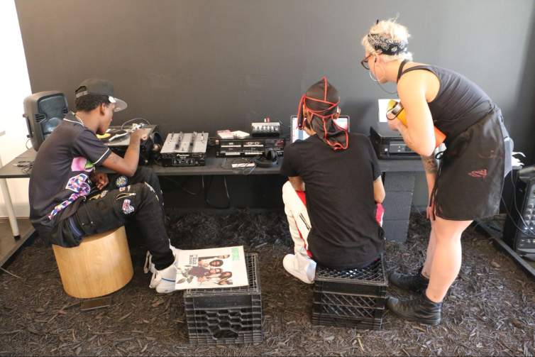 “Disco Gardens” is an interactive sculptural installation, and in this view 3 people are gathered along a low long table outfitted with a turntable, mixers, cassette players, and a laptop. They work with the various equipment, sitting on milk crates or a wood block, and the installation all sits on a bed of brown mulch.