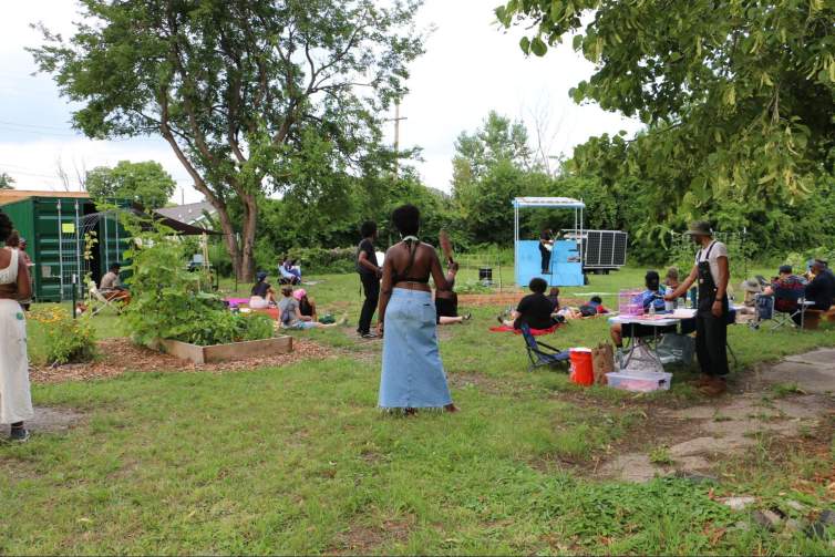 In a grassy park with vegetables grouwing in raised beds, a guitarist plays from a small bright blue solar-powered stage. Onlookers watch, lounging and standing around the lawn.