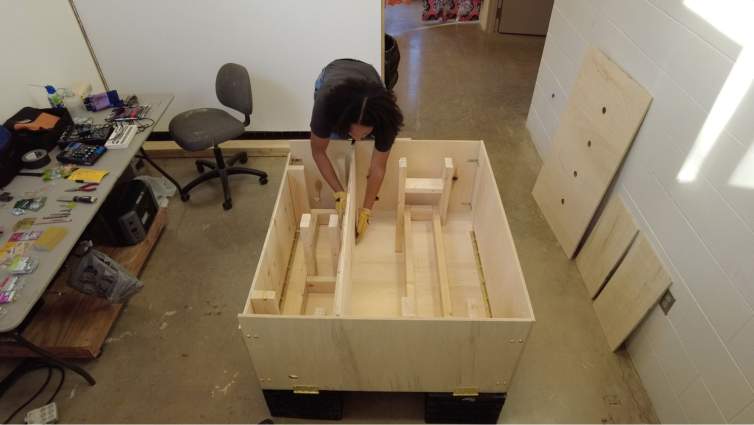 Ash Arder, a Black woman with medium-dark skin tone, leans into a wooden structure that she is building on the floor of a studio space.