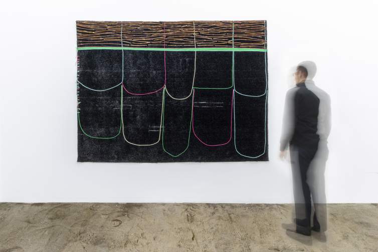A motion-blurred person stands in front of a large black Astroturf rectangle. A thick green stripe marks off the top fourth of the rectangle, with willow sticks swimming horizontally across that area. 10 pieces of multicolor yarn loop down from the top like large irregular shingles.