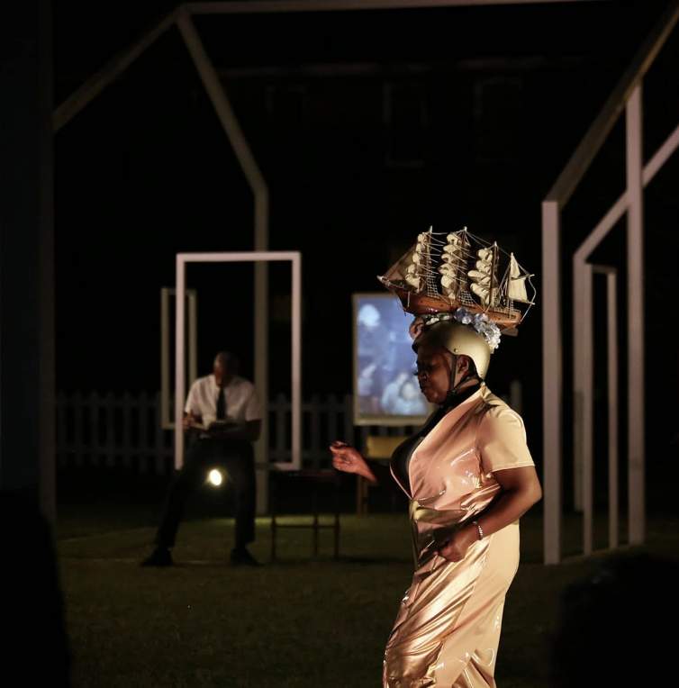 Migratuse Ataraxia is a performance still of the artist wearing a gold helmet with a model sailing ship affixed to the top and a gold and beige jumpsuit. A Black woman, she appears to dance in stride, and a person sits in the darkened background.