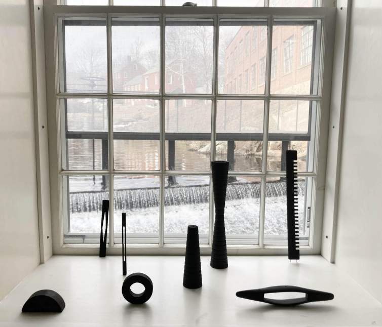 In an artist's studio, eight small black abstract sculptures rest is a deep windowsill. Outside the window, we see rushing water or a river and the side of a brick factory building.