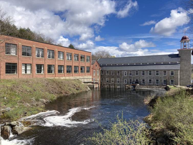 A sprawling brick factory building is perched above a river in a green landscape.