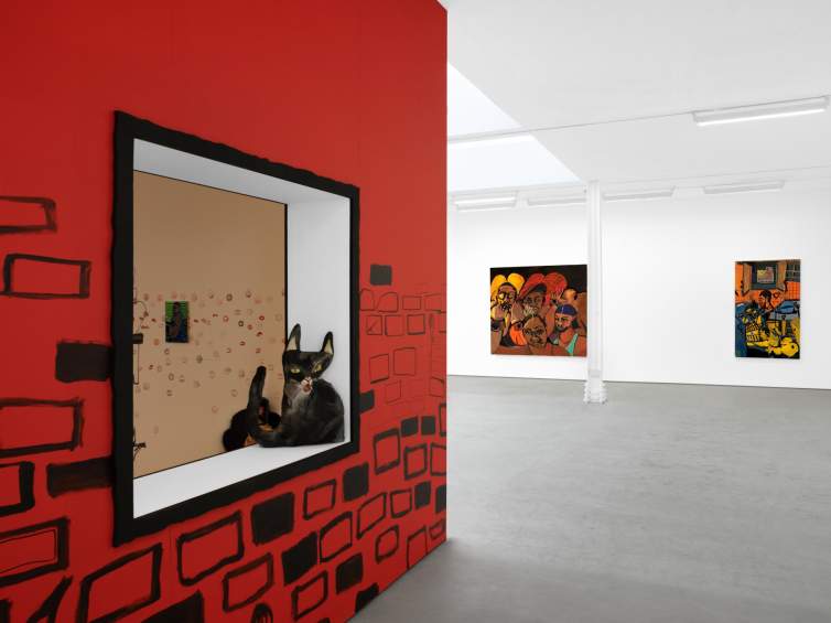 Installed in a white gallery with concrete floors is a large red box with loosely drawn black bricks on the bottom half, with a sewn black cat sculpture with a distorted proportion sitting in a window cut out. On the far wall are two large colorful paintings.