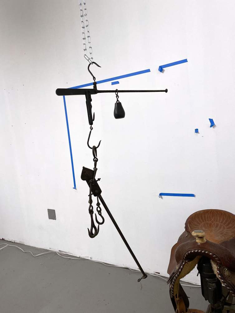 In Peggy Chiang's studio, a black metal mobile composed of connected hooks, weights, and rods hang from the ceiling. we see strips of painter's table on the wall behind and an ornate leather saddle in the corner.
