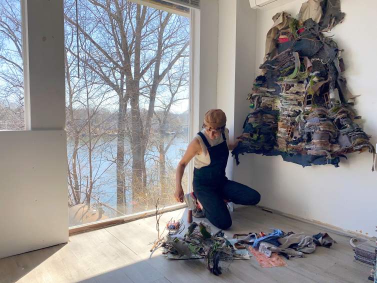 Ruby Chishti kneels in the corner of her studio, a room with white walls and wooden floors, with a window behind her showcasing a view of a lake and trees. Ruby is a Pakistani-American woman with medium light skin and short dark hair. She wears overalls, white tank top, and brown hat and works with fabric strips on a dress-like sculpture that she is assembling on the wall.