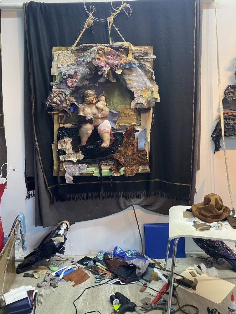 A view of Ruby Chishti's studio shows a work-in-progress with an outer frame and a fabric sculpture handing inside. Various scraps of fabric and art-making materials are strewn about on the floor.