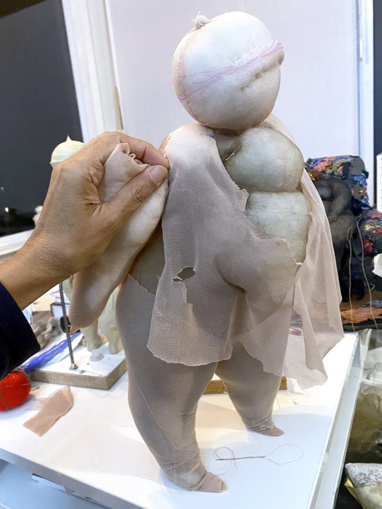 On a work table in Ruby Chishti's studio, a hand with medium skin ton holds a section of stuffed fabric up to a female form created with cotton stuffed into shaped sections of pantyhose.