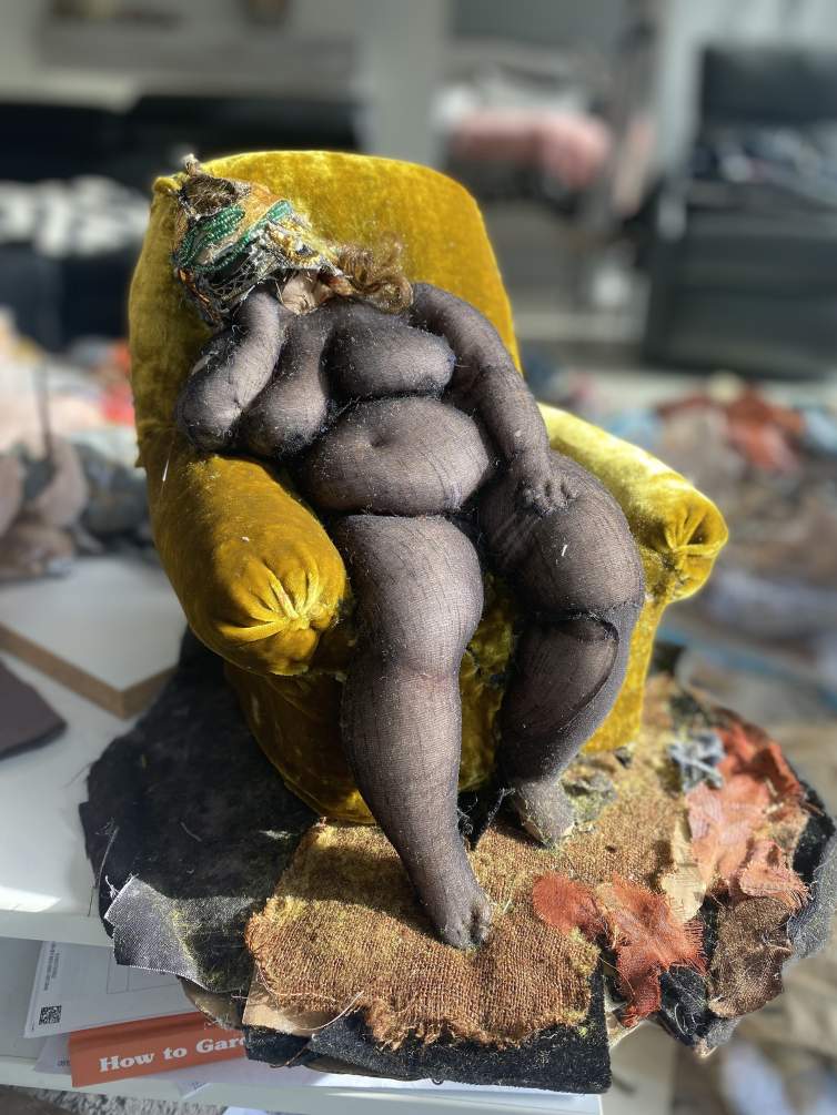 In Ruby Chishti's studio, a soft sculpture of a voluptuous female form sits in a gold velvet armchair with head resting on hand.