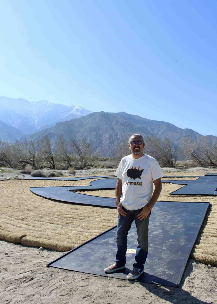 Gerald Clarke stands on a land art installation of a black pathway stretching behind him, installed in Palm Springs desert with  mountains visible in the background under a clear blue sky is a citizen of the Cahuilla Band of Indians, and has a medium skin tone and a salt-and-pepper goatee, and his hair is pulled back, wand wears a tshirt with outline of the US reading “amnesia”.