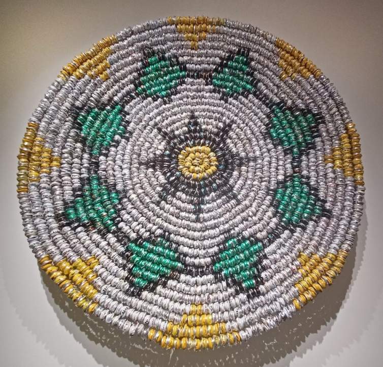 Continuum Basket: Creation is a large, circular wall hanging in the style of Cahuilla coiled basketry, with a geometric pattern in silver, green, black, and yellow formed of crushed cans in concentric circles, displayed on a white wall.