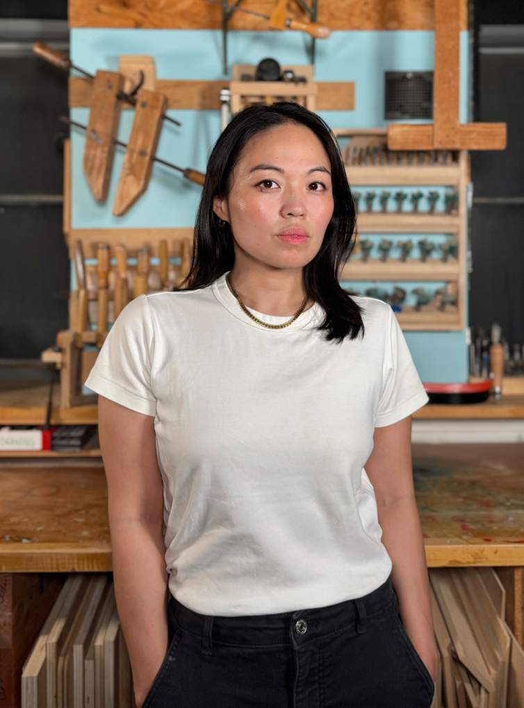 Cathy Della Lucia looks directly at camera in a workshop with clamps and tools on the wall, with her hands in pockets. She is a woman with Korean heritage and has long dark hair. She wears a white t-shirt and dark jeans.