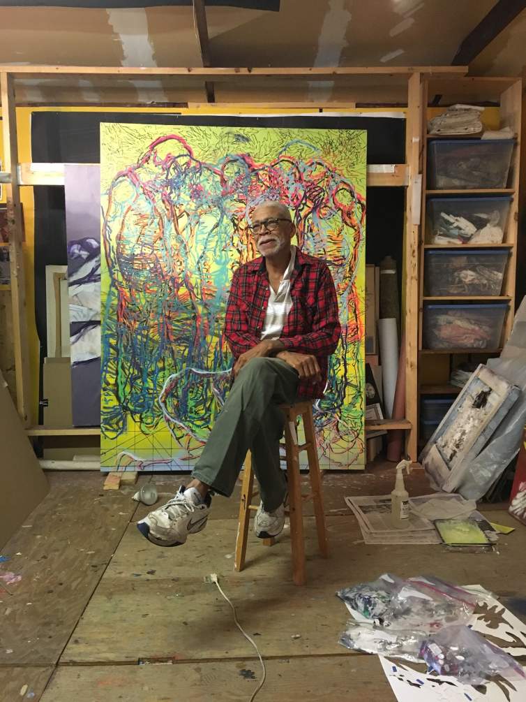 Bob Dilworth sits on a wooden stool in a studio with pine shelves and plywood floors, with a large abstracted figurative painting leaning behind him. He is an older Black man with short-cropped gray hair, and wears a plaid shirt and rounded tortoiseshell glasses.