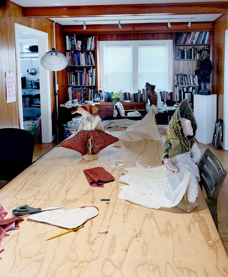 At least three doll-like figures stand on a raw wood work table. Each has a skirt with a train and a high headdress.