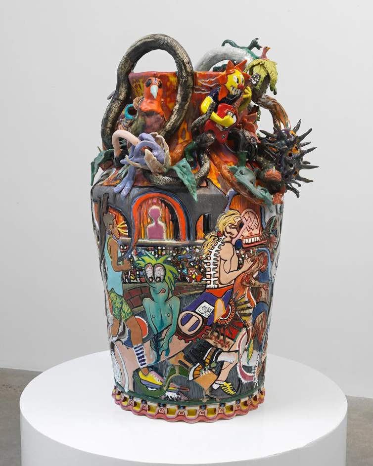 A colorful, eclectic ceramic vase featuring various cartoonish running figures and abstract designs in relief,  with a flower head figure playing electric guitar, a bird and snake affixed to the top, displayed on a white pedestal.