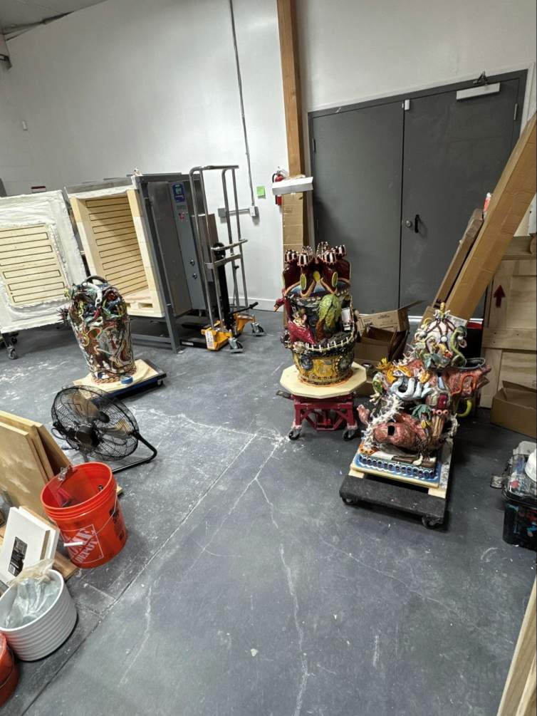Large ceramic sculptures on wheeled platforms in a warehouse room with kilns, fans, and crates nearby.