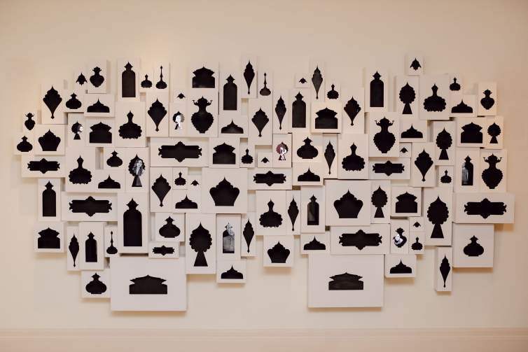 An art installation featuring numerous black silhouette cutouts of various shapes and sizes on white backgrounds, arranged in a grid on a wall. The viewer can peer through some of the shapes to see video imagery inside.