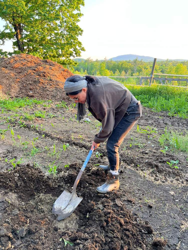 Raheleh Filsoofi, an Iranian-American woman with medium skin tone and dark hair, uses a shovel to collect dirt from a field with fences, trees, and mountains visible behind her. She wears a gray headband, an oversized gray sweater, jeans, work boots, and sunglasses.