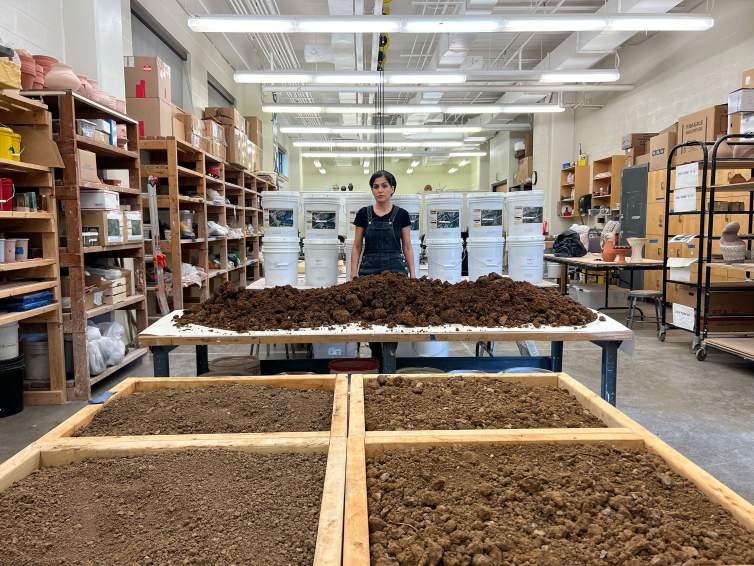 Raheleh Filsoofi, an Iranian-American woman with medium skin tone and dark hair, stands behind two tables full of dirt being processed in her ceramics studio. Behind her are stacks of buckets and the walls are lined with shelves holding various objects and materials.