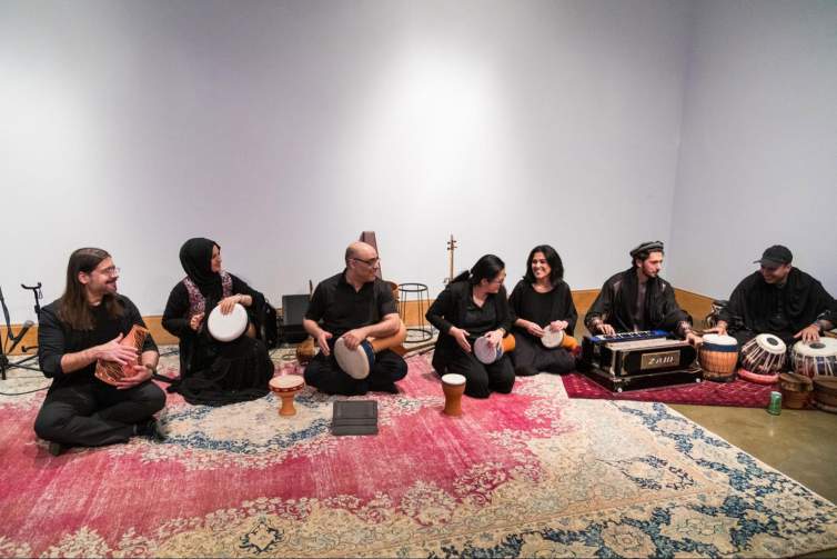 Seven people of different ages and ethnicities, all wearing black, sit on the floor of a white-walled gallery space on a Persian rug, holding handmade clay instruments and other musical equipment. They smiled and laugh with each other while playing the instruments.